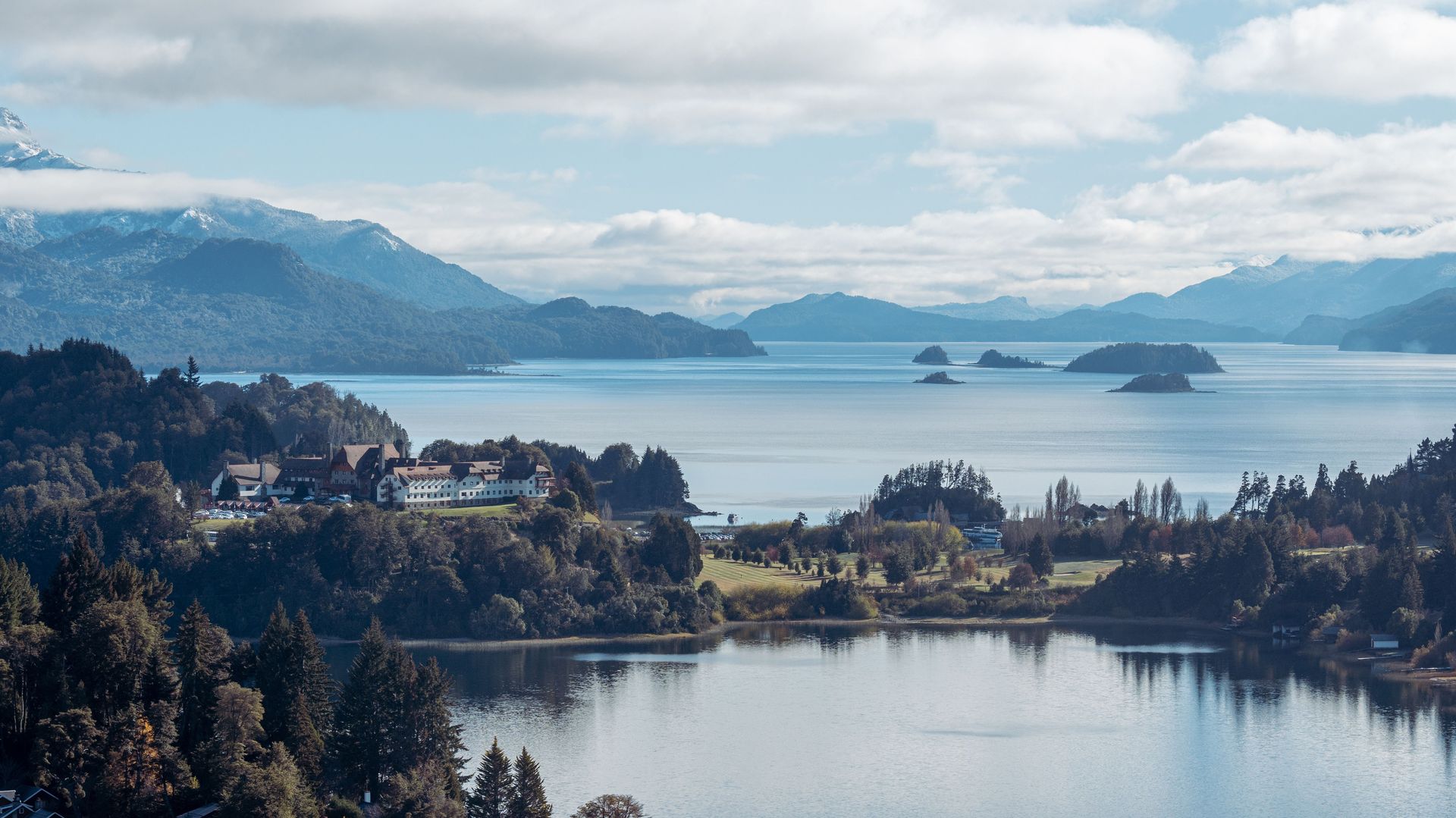 Une vue aérienne époustouflante du lac Nahuel Huapi, avec des forêts luxuriantes et des montagnes lointaines sous un ciel nuageux.